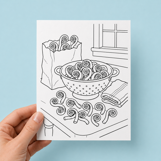 Hand holding a black-and-white colouring greeting card illustrated with fiddleheads piled in a colander, spilling from a paper bag, and scattered across a kitchen table near a window, against a light blue background.
