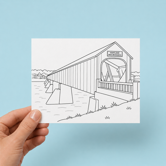 Hand holding a black-and-white colouring greeting card illustrated with the Hartland Covered Bridge in New Brunswick, showing its long wooden span over a river with a village and trees in the background, against a light blue background.