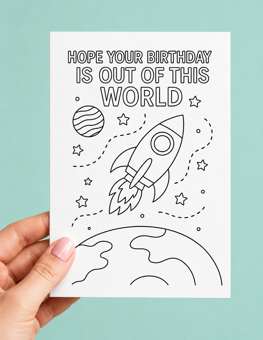 A hand holds a white birthday card featuring a black line art design of a rocket ship, stars, and planets, with the text 'HOPE YOUR BIRTHDAY IS OUT OF THIS WORLD'. The background is a solid light teal color.