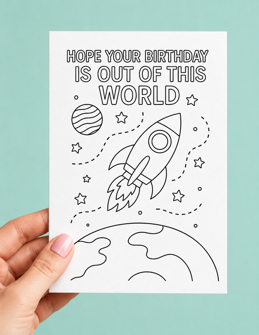 A hand holds a white birthday card featuring a black line art design of a rocket ship, stars, and planets, with the text 'HOPE YOUR BIRTHDAY IS OUT OF THIS WORLD'. The background is a solid light teal color.